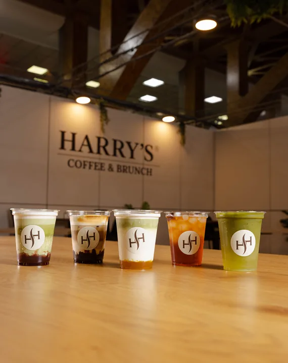 Cinco bebidas de Harry's Coffee & Brunch alineadas sobre una mesa de madera frente al logo en la pared