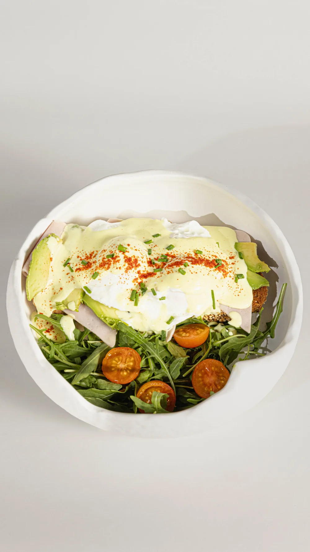 Tosta Benedict con huevo poché, aguacate, jamón y salsa holandesa en bowl blanco con rugula y tomates