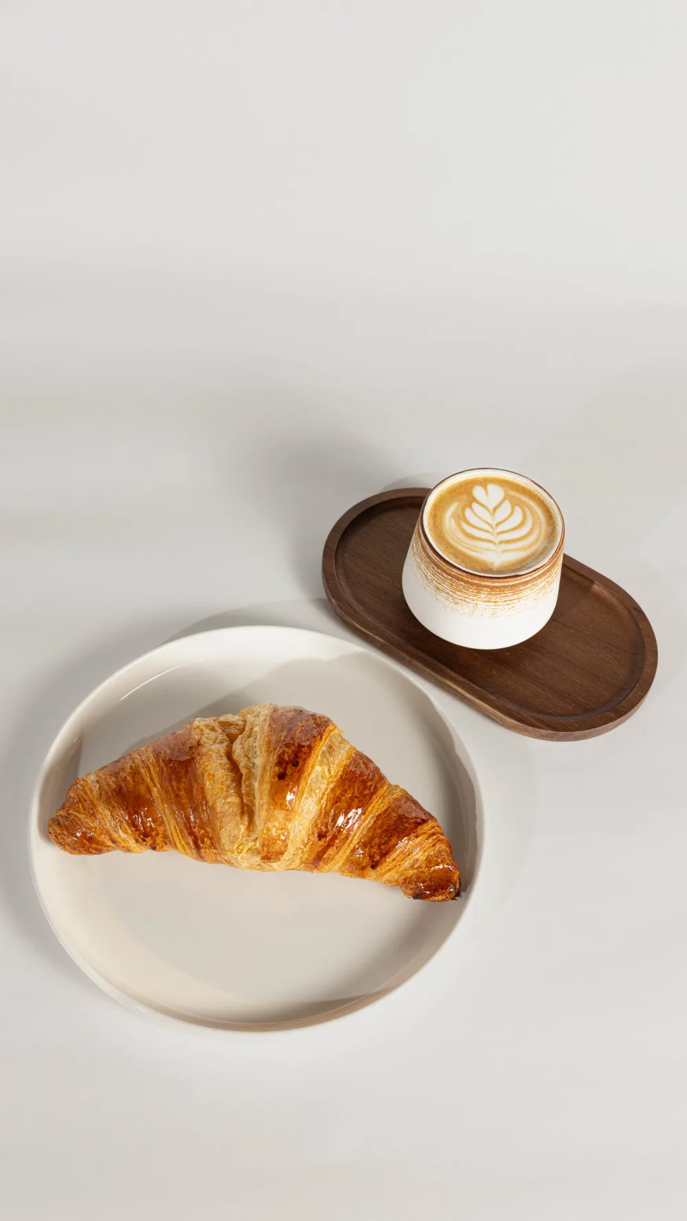 Croissant recién horneado en plato cerámico junto a latte con arte de hoja en taza sobre bandeja de madera