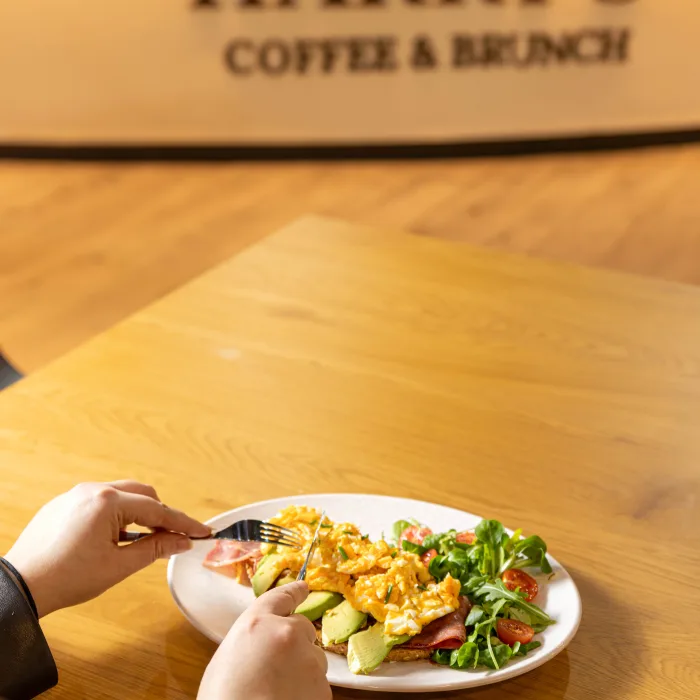 Manos cortando tosta de aguacate con huevo revuelto en mesa de madera bajo el logo de Harry's Coffee & Brunch